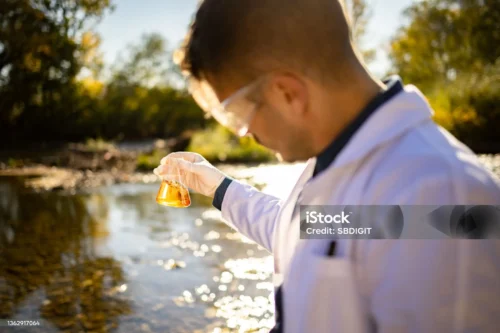 A person in a lab coat and protectivce goggles studies coloured liquid in a small chemical flask, beside a beautiful river.